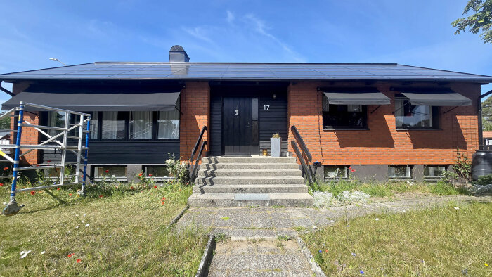 Tegelhus med tre svarta markiser installerade, byggnadsställning syns till vänster, grön gräsmatta och blå himmel i bakgrunden.