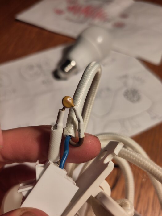En hand håller en vit elkabel med en liten komponent täckt av krympslang, bredvid syns en LED-ljuskälla på ett bord med papper i bakgrunden.