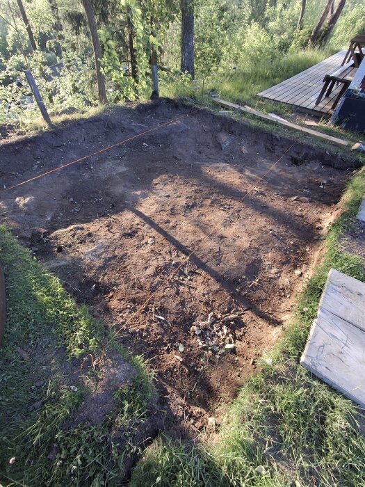 Utgrävd grop för växthusbygge med två spända snören i olika höjder för nivåkontroll, omgiven av gräs och träd.