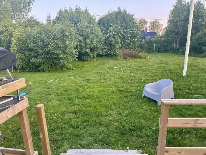 Gräsmatta med en blå plaststol och altanräcke, omgiven av buskar och träd under en ljus himmel.