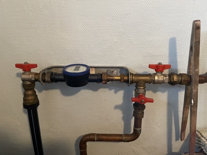 Vattenmätare med anslutna rör och röda ventiler monterade på en vit vägg. Ett förlängningsrör syns till höger om mätaren.