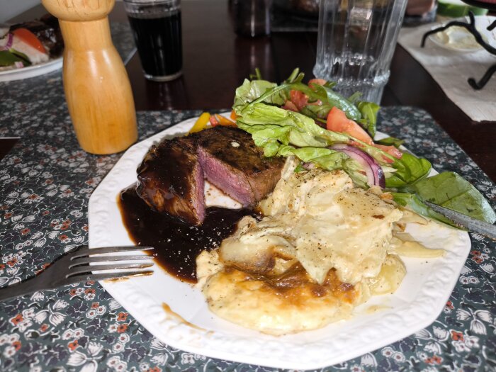 Tallrik med grillat kött, potatisgratäng och sallad, samt en pepparkvarn och dryck i bakgrunden.