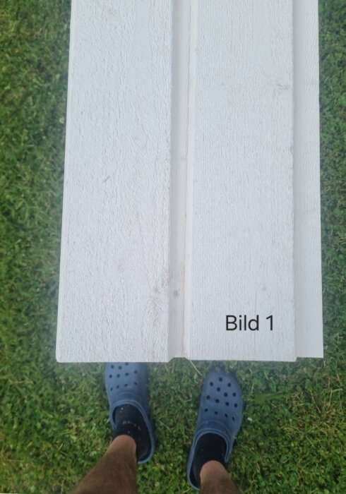 Vertikalt monterad vit baspanel på gräsmatta, med ben och fötter i tofflor synliga nedanför panelen.