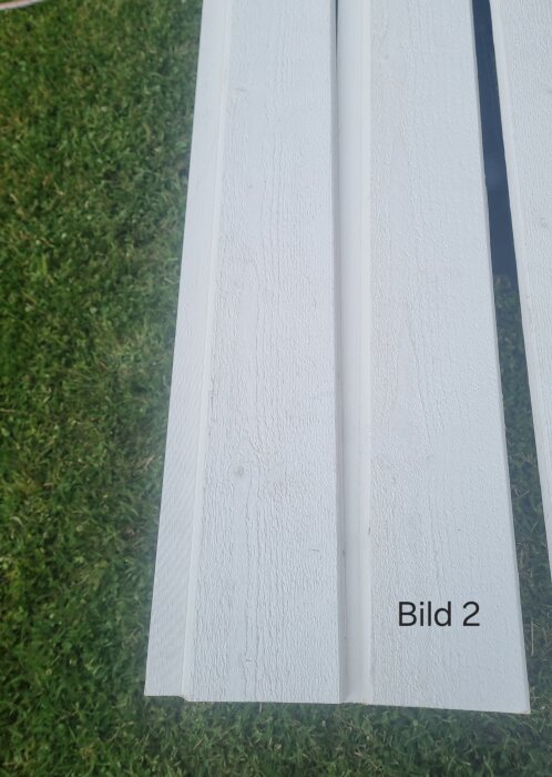 Vitmålad träpanel stående på gräs, märkt "Bild 2". Brädorna har tydlig trästruktur och är avsedda för montage som baspanel.