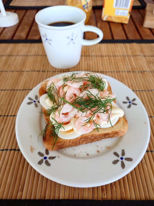 Räksmörgås på en blommig tallrik med dill och ägg, kopp kaffe i bakgrunden på en bambumatta.