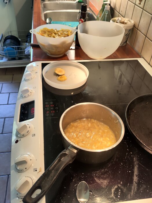En kastrull med citronmarmelad på en spis, omgiven av skålar med citronskal och en sked, i ett kök under provkok av flädermarmelad.