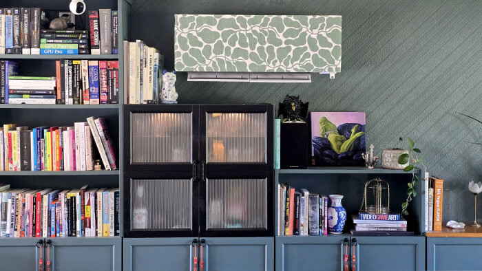 Väggmonterad luftvärmepump täckt av grönmönstrad tapet, omgiven av bokhyllor med böcker och dekorativa föremål i ett vardagsrum.