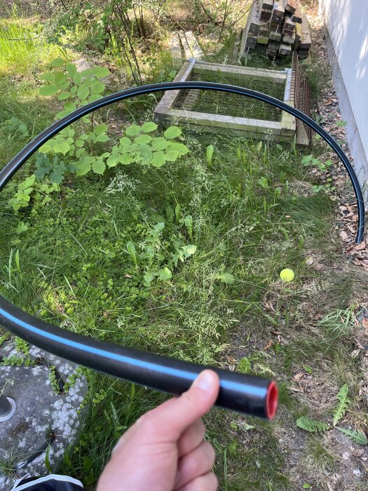Person håller en svart PEM slang med blå rand i en trädgård vid sidan av ett hus, omgiven av gräs och träd.
