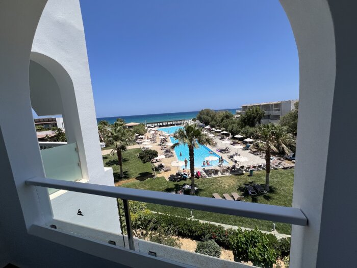 Utsikt från en balkong över en pool, grönområde och hav, med solstolar och parasoller runt poolen.