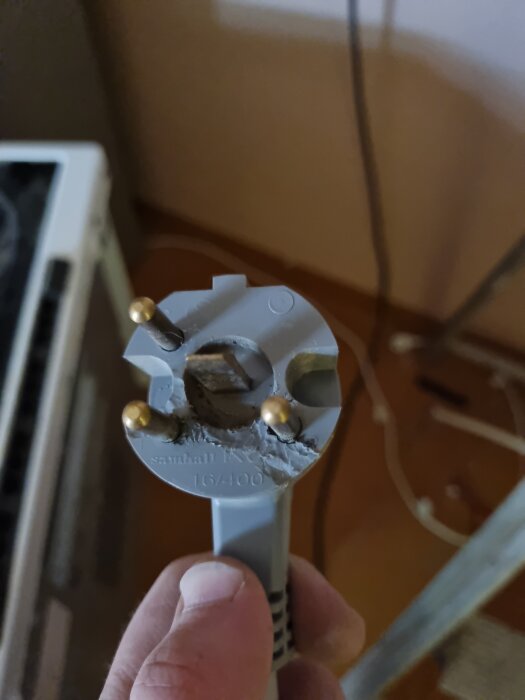 Trepoligt spisuttag med skador på kontakten i närbild, hålls av en hand, del av diskussion om elinstallation för en gammal spis.