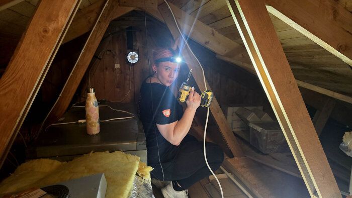 Person med pannlampa arbetar på vinden med ett verktyg för kabeldragning, omgiven av träbalkar och verktyg.