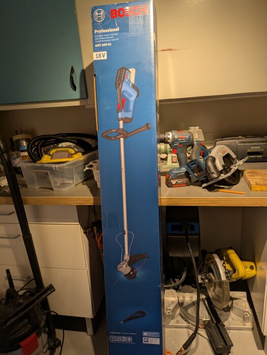 Förpackning till en Bosch batteridriven trimmer, 18V, placerad i ett garage med verktyg i bakgrunden.