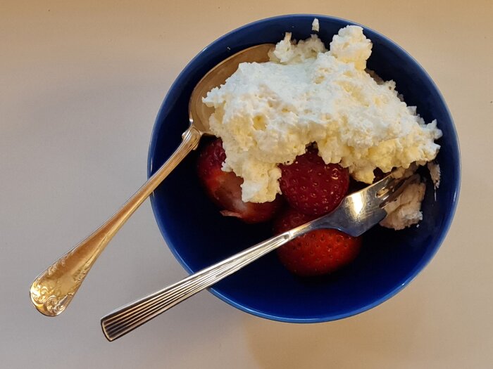 Blå skål med jordgubbar och vispad grädde, serverad med en sked och gaffel.