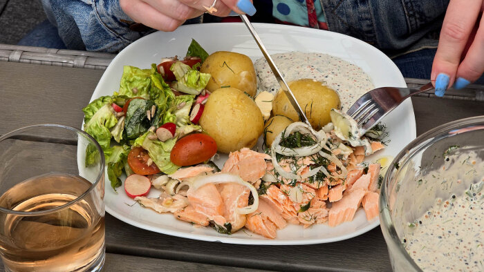 Tallrik med lax, potatis, sallad och sås. En hand med blått nagellack håller bestick. Glas med dryck bredvid tallriken.
