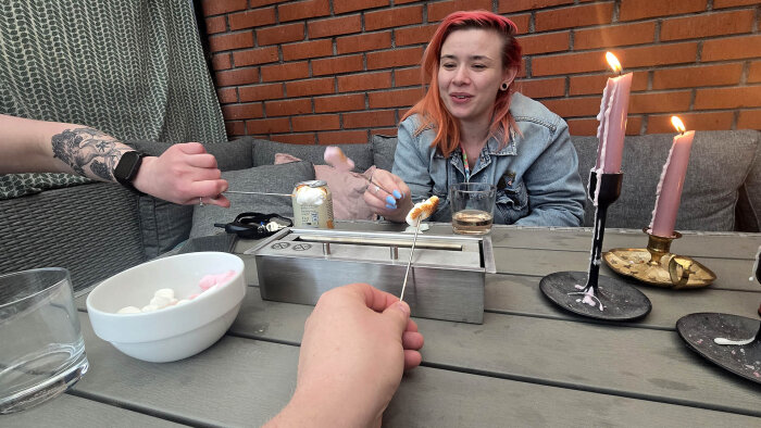 Personer grillar marshmallows vid ett bord med ett litet bordsmatlagare, rosa ljus och skålar på bordet.