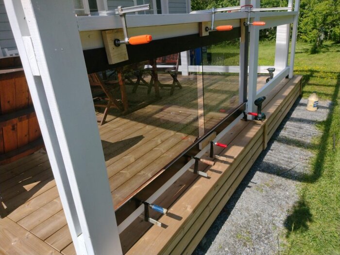 Glasmontering på altan med trästaket, fäst med klämmor. Pågående projekt för att anpassa glas i räcket. Verktyg synliga på trallen.