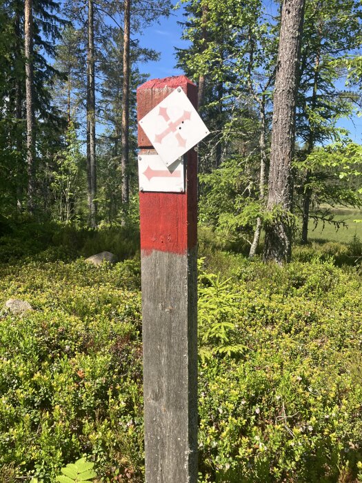 Röd trästolpe med två vita skyltar i skogsmiljö, omgiven av gröna buskar och tallar under en blå himmel.
