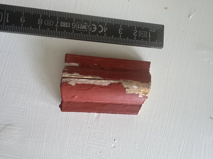 Rött målat trästycke med avflagad färg och skador, bredvid en linjal som visar mått i centimeter.