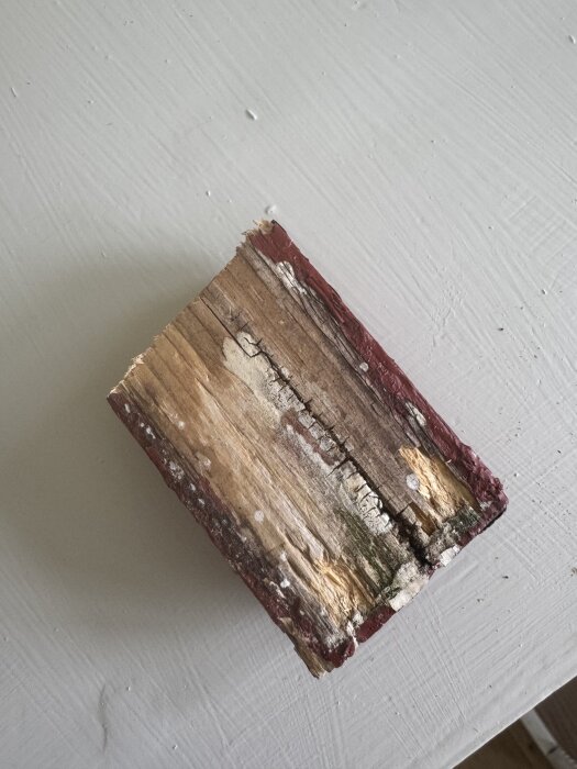 Ruttet trästycke med avskavd röd färg visar skador och potentiellt behov av byte, liggande på ett vitt underlag.