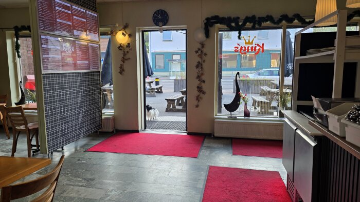 Hund vid öppen dörr i pizzeria, inomhusmiljö med röda mattor och dekorationer. Ute finns picknickbord och gatuskylt.