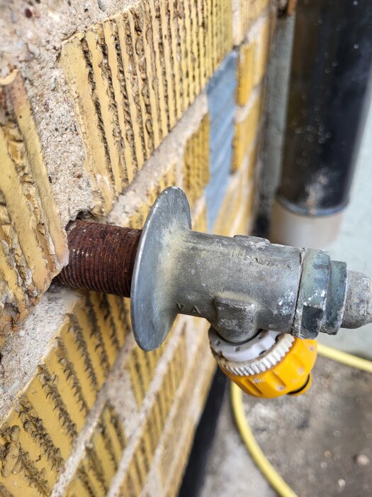 Utvändig vattenkran med rostigt rör fäst i gul tegelvägg, lågtrycksproblem efter lång oanvändning, kräver eventuell demontering för inspektion.