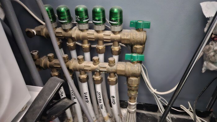 Rörsystem med kopplade vattenventiler, märkta "VARD", "STORA SOV", och "BAD". Gröna vred på ventilerna i närbild.