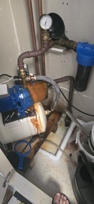 En vattenpump med synlig rost och läckage vid en anslutning, stående på ett golv. Slangar och rör kopplade till pumpen.