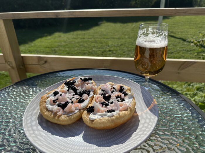 Langos toppad med räkor, lök och svart kaviar på en tallrik bredvid ett glas öl, utomhus på ett glasbord.