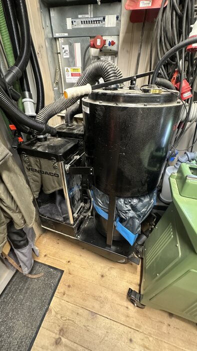 Grovsug med cyklon och dammsugarmotorer i snickerboa, omgiven av slangar och utrustning, på trägolv.