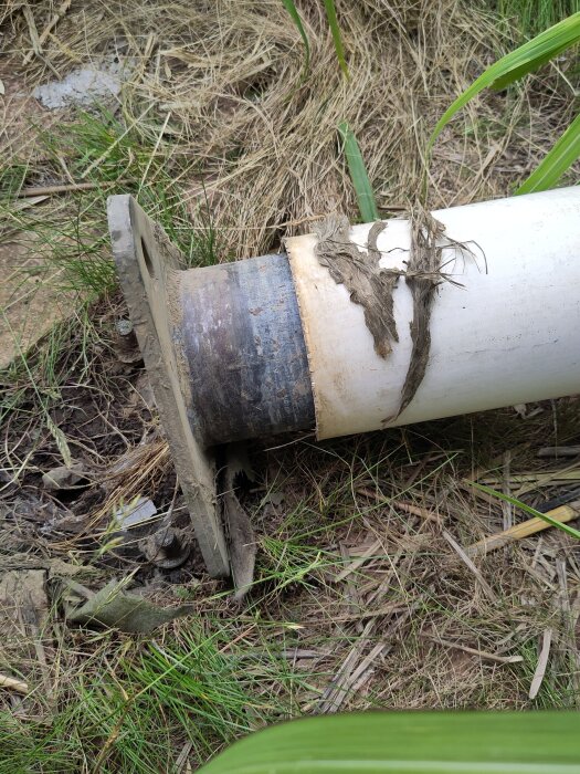 Flaggstång på marken med skadad bas, omgiven av gräs. Material, tidigare inuti, ligger löst på stången.