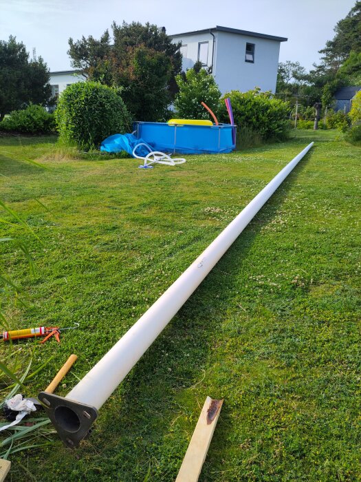 Vit flaggstång ligger på gräsmatta nära en blå pool och ett modernt hus i bakgrunden.