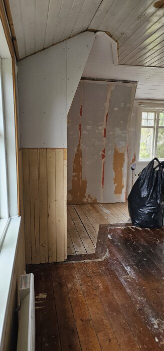 Rum under renovering i ett hus med exponerade träväggar och tak. Plastpåse och fönster synliga i bakgrunden.