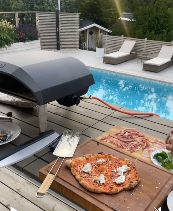 En nygräddad pizza med ost på en skärbräda vid en utomhusugn nära en pool, med solstolar i bakgrunden.
