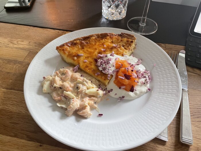 Tallrik med bit av matpaj, rom, rödlök och kräftröra, på ett träbord med glas och bestick.