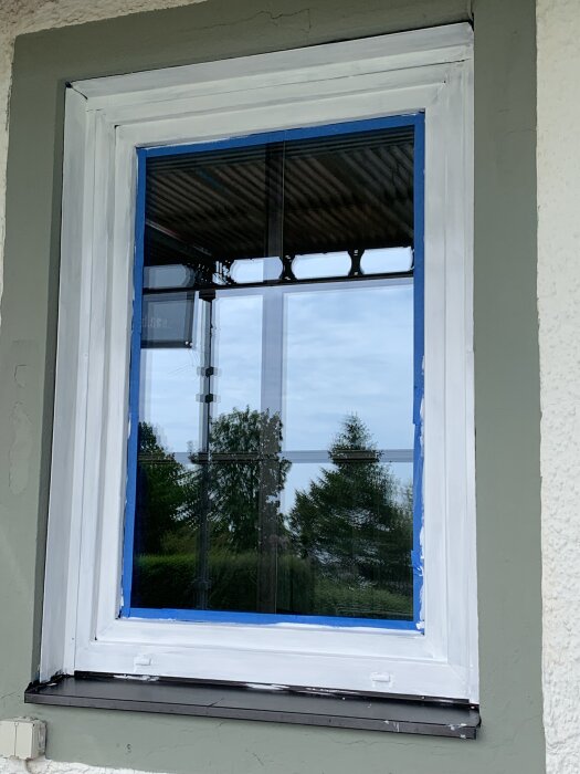 Fönster med svarta spröjsar mellan rutorna, inramat av vit färg och blå tejp, omgiven av husets vägg.