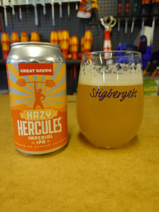 En ölburk med etiketten "Hazy Hercules Imperial IPA" och ett glas fyllt med öl på ett bord. Bakgrunden är en verkstad med verktyg.