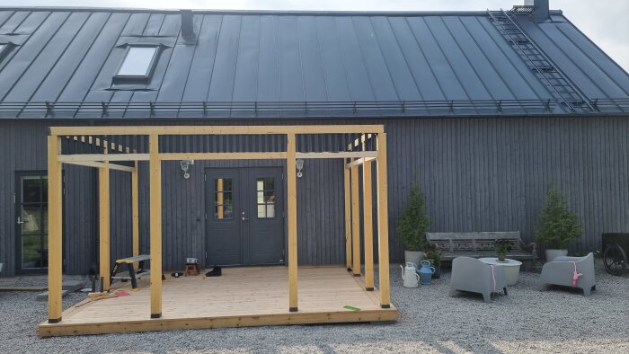En träpergola under konstruktion framför en grå byggnad med ett plåttak. Stolar och trädgårdsdekorationer syns i förgrunden.