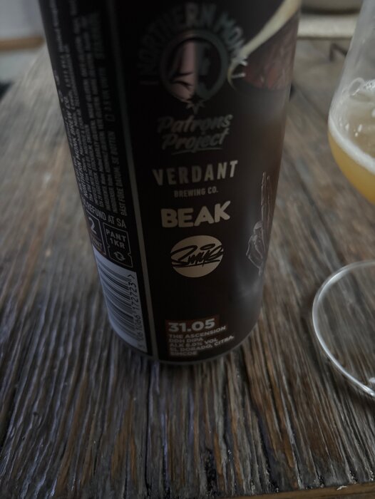 Burk med Northern Monk och Verdant Patrons Project öl, bredvid ett glas med öl på ett träbord.