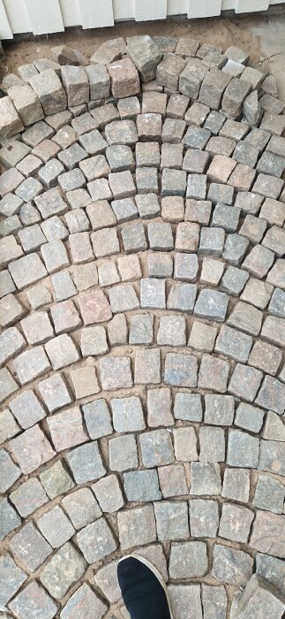 Smågatsten lagda i ett cirkulärt mönster på en grusad uppfart, med en vit träfasad i bakgrunden och en sko synlig i hörnet.