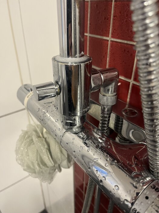 Kromfärgad duscharmatur med vattenläckage vid reglaget mot röd kakelbakgrund.