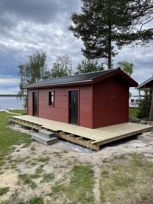 Röd stuga vid sjö med ny altan under konstruktion, saknar två trallbrädor.