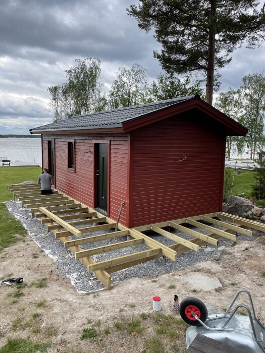 Röd stuga vid sjön med påbörjad altan av träreglar, byggd under midsommarhelgen. En skottkärra och byggmaterial syns i förgrunden.