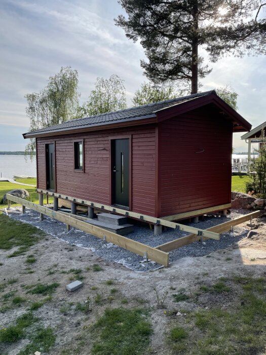 Altanbygge vid rött fritidshus nära sjön, med träram monterad men saknar två längder av trallbrädor, omgiven av grus och gröna träd.