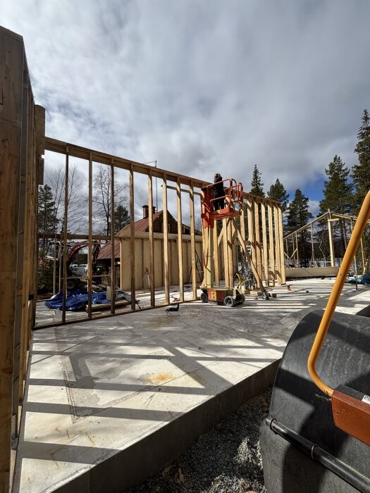 Byggarbetsplats med stomme till vägg under uppförande, en person i lift arbetar på stommen, molnigt väder.