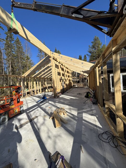 Pågående takbygge med trätakstolar under montering, fint väder och byggmaterial utspritt på betonggolv. Skog i bakgrunden.