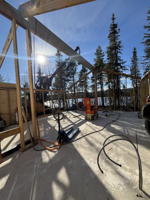 Bygge av husstomme under klarblå himmel, med ett kranfordon lyftandes takbjälke, omgivet av barrträd. Solen skiner genom trästrukturen.