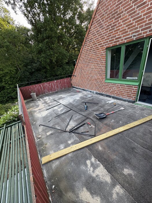 Balkong med tillbyggnadstak, verktyg och material på ytan som ska renoveras, mur av tegel och omgivande grönska.
