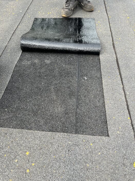 Takrenovering med asfaltpapp, delvis avtäckt underlag och synliga arbetsskor i bildens ovankant.