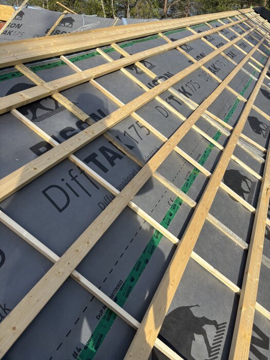 Byggprojekt med trätakstolar och takpapp, där dubbelhäftande tejp syns vid övergången mellan vägg och tak för ökad stabilitet.