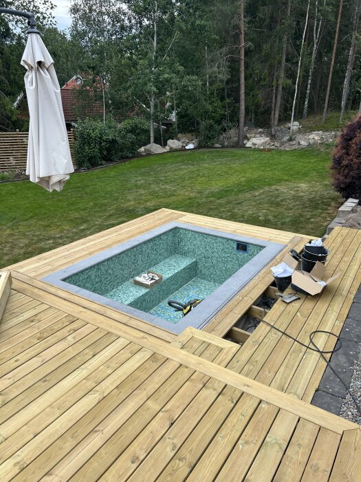 Trädäck med pool under konstruktion, omgiven av grönskande gräsmatta och skog. Material och verktyg ligger utspridda på trädäcket.
